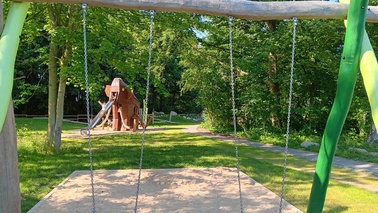 Im Hintergrund stehender Kletterturm im Stil eines Mammuts mit Rutsche als Rüssel, auf der Rutsche ein fröhliches Kind beim Rutschen, im Vordergrund steht eine Schaukel