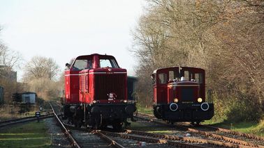 Two historic locomotives ride on tracks through a forest area.